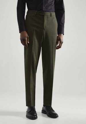 Homme portant un pantalon sur mesure vert olive associé à des chaussures habillées noires et une chemise noire boutonnée, debout devant un fond uni.
