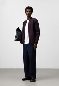 Bordowy sweter, biały t-shirt i granatowe spodnie. Trzyma czarną skórzaną torbę. Gładka tekstura, zamek z przodu, luźny krój, minimalistyczny design.