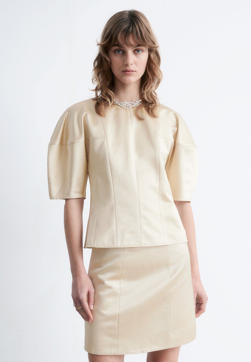 Cremefarbenes Kunstlederoberteil mit Puffärmeln und strukturierter Passform, kombiniert mit einem passenden Minirock. Verfügt über einen dezenten Glanz und klare Linien.