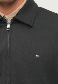 Crna jakna sa zatvaračem i šiljastim ovratnikom, izrađena od teksturirane tkanine, s malim crveno-bijelim-plavim logotipom na lijevoj strani prsnog koša.