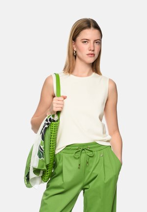 Woman with shoulder-length blonde hair wearing sleeveless white top, green drawstring pants, holding green woven bag with patterned scarf.