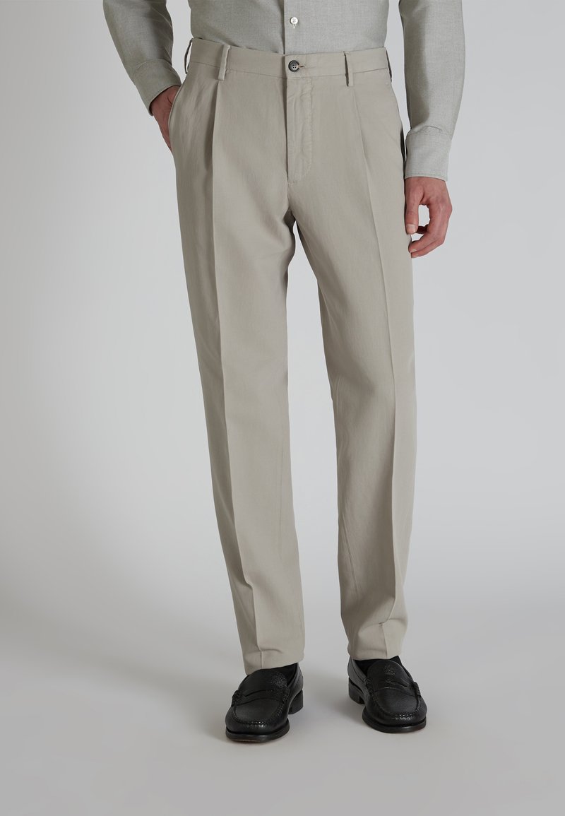 Pantalones plisados beige con cremallera, cierre de un botón y bolsillos laterales; hechos de una tela suave y ligera. Se muestran zapatos negros.