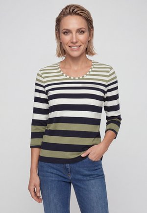 Frau mit kurzen blonden Haaren, die ein grün-weiß-schwarz gestreiftes Langarmshirt und blaue Jeans trägt, lächelnd mit einer Hand in der Hosentasche.