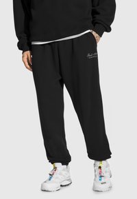 Czarne dresy z elastycznymi mankietami, z małym białym logo na lewym biodrze. Noszone z białymi butami sportowymi z kolorowymi akcentami.