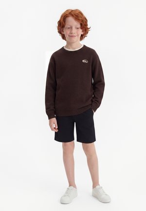 Rødhåret dreng iført en mørkebrun sweater, sorte shorts og hvide sneakers står med den ene hånd i lommen, smilende let, på en hvid baggrund.