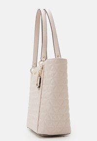 Lichtroze doorgestikte shopper met twee lange hengsels, gouden accenten en een decoratieve hanger aan de zijkant. Gewelfde oppervlakdetail.