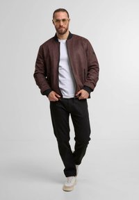 Jachetă bomber din suede maro, cu guler și manșete ribbate negre, închidere cu fermoar în față și două buzunare laterale, purtată cu pantaloni negri și adidași albi.