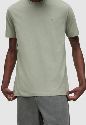 Homme portant un t-shirt vert clair à manches courtes et un short gris, tenant l'ourlet du bas du t-shirt avec les deux mains.