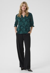 Blusa floreale verde con scollo a V e maniche trasparenti a tre quarti, abbinata a pantaloni neri a gamba larga e décolleté neri a punta.