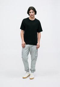 Ung man står i en svart t-shirt, ljusgrå cargobyxor och vita sneakers med gummisulor mot en enkel vit bakgrund.