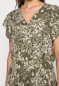 Zelené květinové šaty s výstřihem do V, krátkými rukávy a nařaseným pasem. Tyto šaty mají bílé květinové vzory na tmavě zelené texturované látce.