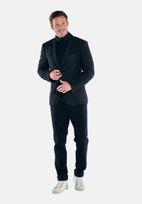 Marineblazer aus strukturiertem Stoff, passender Rollkragenpullover, dunkle Hose und weiße Sneakers. Das Outfit weist moderne Schneiderkunst und klassische Akzente auf.