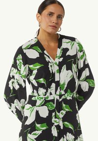 Blumenkleid mit schwarzem Hintergrund, ausgestattet mit großen weißen und grünen Blattmustern, einem tiefen V-Ausschnitt und gefalteten Details an der Vorderseite.