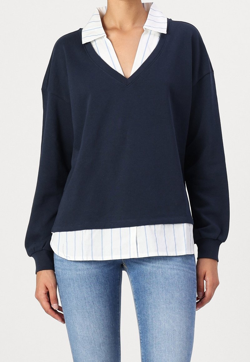 Sweat-shirt bleu marine à col en V en matière douce, porté sur une chemise blanche à rayures verticales bleu clair, avec des manches amples.