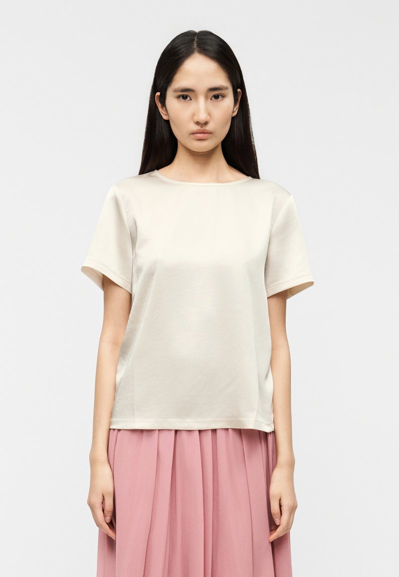 Κρεμ τοπ με κοντά μανίκια και στρογγυλή λαιμόκοψ η, με λεία υφή και άνετη εφαρμογή, σε συνδυασμό με μια ροζ πλισέ φούστα από κάτω.