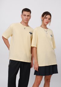 Geltoni medvilniniai didelio dydžio marškiniai su trumpomis rankovėmis, puikuojantys grafikiniu logotipu. Vienas modelis derina su juodomis sportinėmis kelnėmis, kitas - su plisuota sijonu.