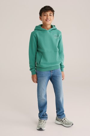 Sudadera verde con capucha, bolsillo frontal y puños de canalé, combinada con jeans azul claro y zapatillas grises con acentos negros.