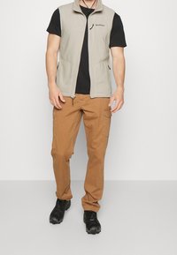 Beige fleeceväst med hög krage, svart t-shirt under, beige cargobyxor och svarta skor. Framfickor på västen för förvaring.