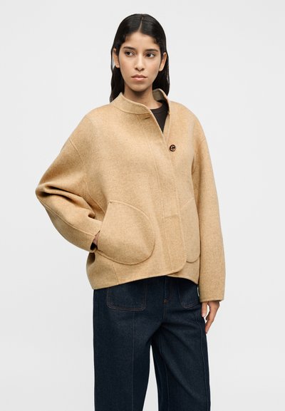 Μπεζ σακάκι από μείγμα μαλλιού με ψηλό γιακά, κλείσιμο με κουμπιά μπροστά και δύο μεγάλες μπροστινές τσέπες. Στρογγυλεμένο σχήμα με άνετη εφαρμογή.