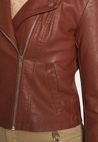 Brązowa skórzana kurtka motocyklowa z srebrnym zamkiem, skośną kieszenią na klatce piersiowej i kołnierzem na zatrzask, noszona na beżowym swetrze dzianinowym i khaki spodniach.