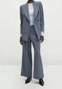 Grijze op maat gemaakte blazer met scherpe revers over een lichtblauwe top, gecombineerd met brede grijze broeken en slanke zwarte hakken. Soepele stofstructuur.