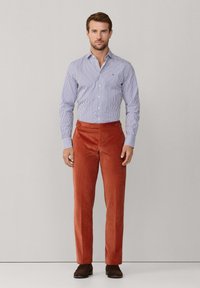 Man standing wearing a blue and white striped button-up shirt, rust-colored corduroy pants, and dark brown shoes against a plain background.