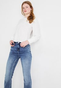 Top de punto blanco con ojales, de manga larga y cuello redondo, combinado con jeans de mezclilla de color azul claro. Ajustado al cuerpo con una textura y detalles sutiles.