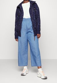 Felpa con cappuccio blu navy con piccoli motivi multicolori, t-shirt bianca, pantaloni larghi in denim chiaro e sneakers grigie con accenti arancioni.