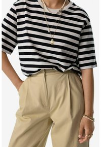 Chemise à rayures noires et blanches à manches courtes avec une coupe décontractée. Associée à un pantalon taille haute beige. Colliers en or à plusieurs couches avec des pendentifs.