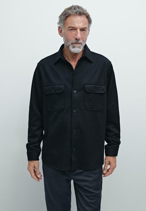 Chemise noire à boutons avec deux poches poitrine, fabriquée à partir d'un tissu texturé. Dotée d'un col et de manches longues. Design simple et moderne.