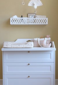 Previjalna mizica z belo lesenim zaključkom, na vrhu bež oblazinjena previjalna podloga, shranjevalna torba in mehka igrača. Nad njo je okrasna polica.