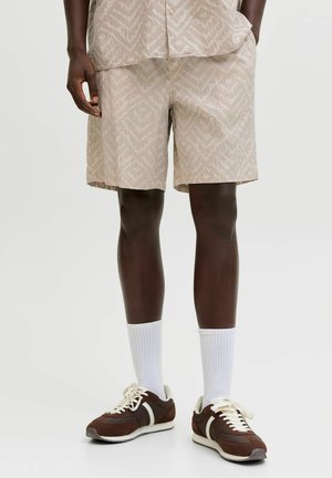 Mann har på seg beige mønstrete shorts, hvite sokker og brune joggesko med hvite lisser og striper mot en lys bakgrunn.