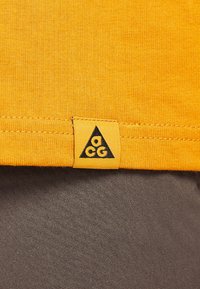 Material de bumbac galben cu o finisare texturată, având o aplicatie triangulară neagră cu branding-ul "ACG" la margine. Material gri dedesubt.