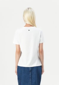 Tricou alb cu mânecă scurtă, decolteu rotund și un mic detaliu logo negru la spate. Textură a materialului moale. Asociat cu o fustă din denim.