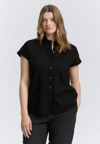 Chemise noire à manches courtes, à boutons, avec un col et des boutons noirs. Le tissu semble lisse et légèrement texturé.