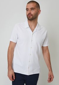 Camisa blanca de manga corta con cuello y una textura suave, con un cierre de botones en la parte delantera y un dobladillo recto, usada con pantalones oscuros.