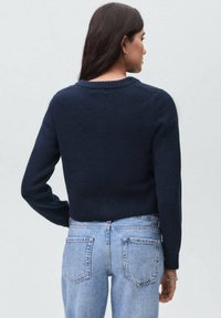 Kvinna med långt mörkt hår iförd en marinblå tröja och ljusblå högmidjade jeans, vänd bort mot en slät grå bakgrund.