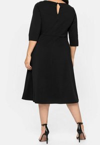 Robe noire avec des manches trois-quarts et une couture à la taille, présentant un détail en forme de trou de serrure à l'arrière. Tissu lisse, jupe évasée, associée à des talons noirs.