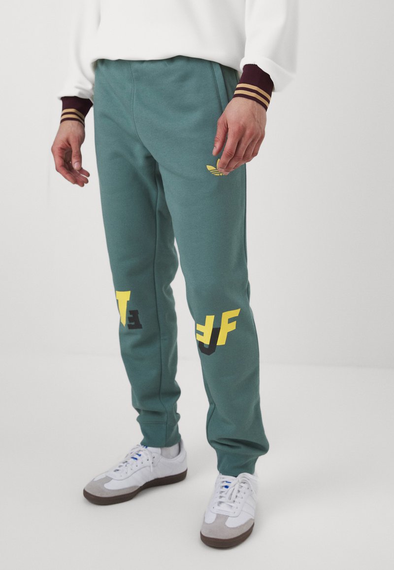 Teal sweatpants with a smooth texture, featuring a yellow logo and bold lettering in black and yellow. Accented with a white and gray sneaker.