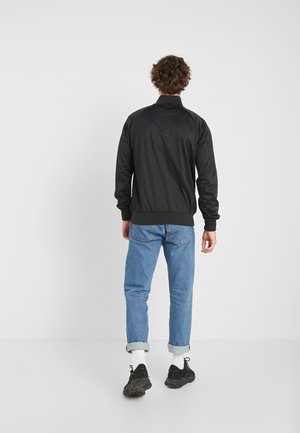 Homme portant une veste noire, un jean bleu clair retroussé, des chaussettes blanches et des baskets noires, debout de dos devant un fond clair et uni.