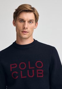 Marinblå stickad tröja med röd "POLO CLUB" text, ribbad rund halsringning och textilstruktur, designad för en kroppsnära passform.