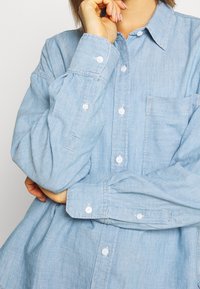 Person i en ljusblå skjorta med knappar och långa ärmar, med ena armen i kors och den andra handen vilande på hakan.