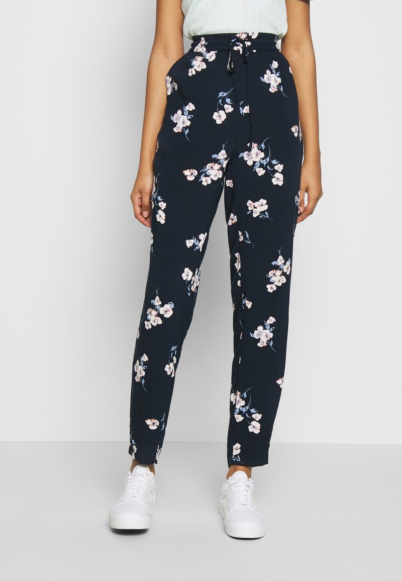 Pantalon marine à fleurs avec taille à cordon, présentant un motif de fleurs roses et blanches et des jambes fuselées, associé à des baskets blanches.