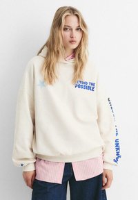Junge Frau mit langen blonden Haaren, die einen übergroßen cremefarbenen Hoodie mit blauer Schrift und einem pink gestreiften Shirt darunter trägt.