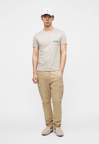 Γκρι κοντομάνικο μπλουζάκι με τσέπη, καφέ cargo παντελόνι με πλαϊνές τσέπες και μπεζ παπούτσια slip-on, ολοκληρωμένο με λευκό καπέλο του μπέιζμπολ.