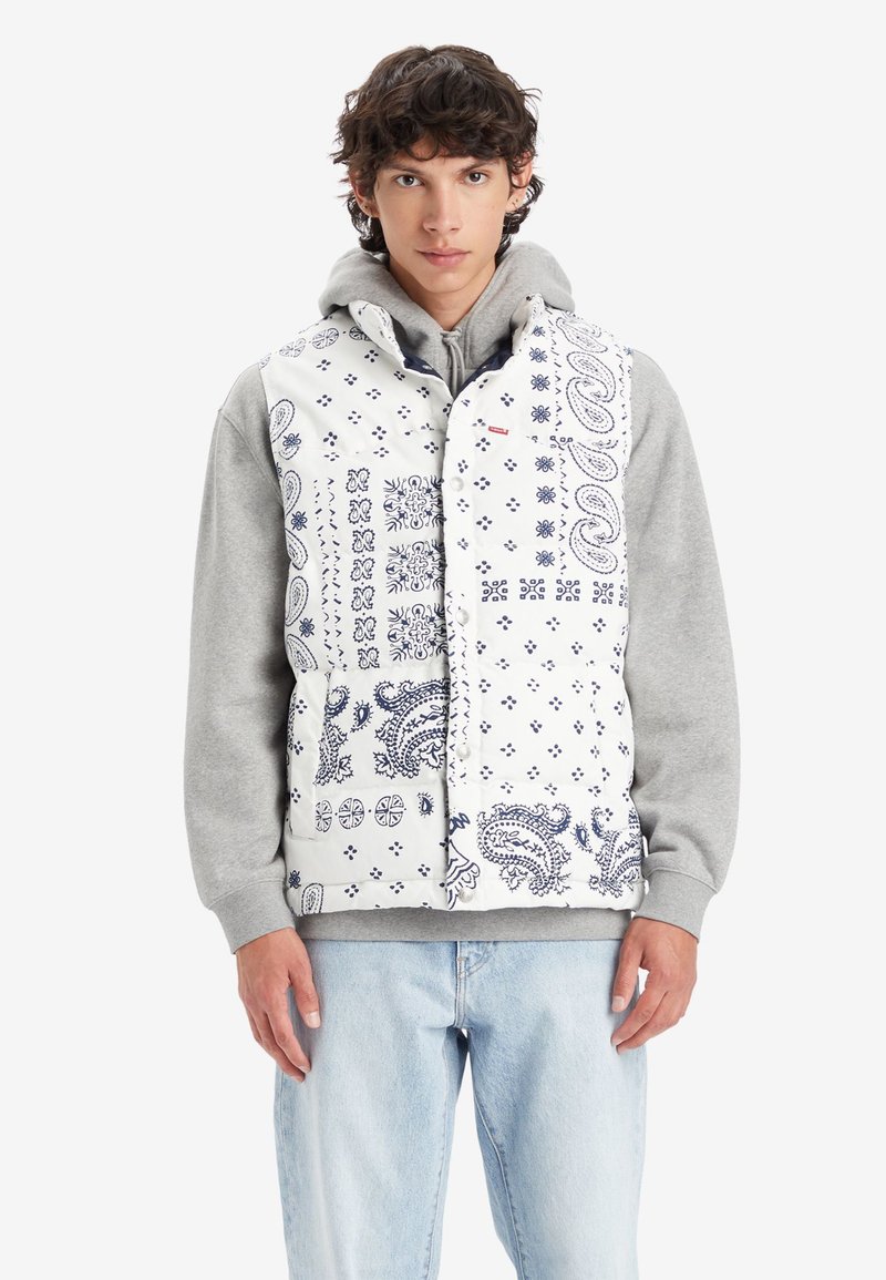 Chaleco acolchado blanco con patrones paisley azules y diseños geométricos, con cierre de botones a presión, superpuesto a una sudadera gris.