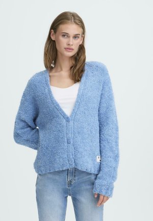 Jeune femme portant un cardigan à boutons texturé bleu clair sur un haut blanc et un jean bleu clair, debout contre un arrière-plan uni.