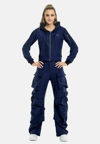 Marinblå hoodie med dragkedja och sidofickor, kombinerat med cargo-byxor med rynkade fickor. Båda är tillverkade av mjukt, stretchigt tyg.