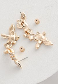 Ohrstecker aus goldfarbenem Metall mit floraler Gestaltung, strukturierten Blütenblättern, einem Kristallakzent und Steckverschluss. Kleine goldene Perlen sind enthalten.