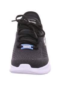 Baskets de sport noires avec tige en maille, lacets plats, semelle blanche et étiquette "LAVABLE EN MACHINE" en bleu. Présente une texture lisse et un design à bout rond.